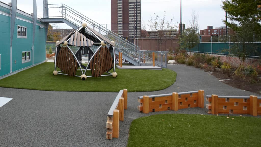 Playground featuring a climbing structure and a slide, surrounded by green grass and pathways, with urban buildings in the background. Ideal for children's outdoor activities and play.