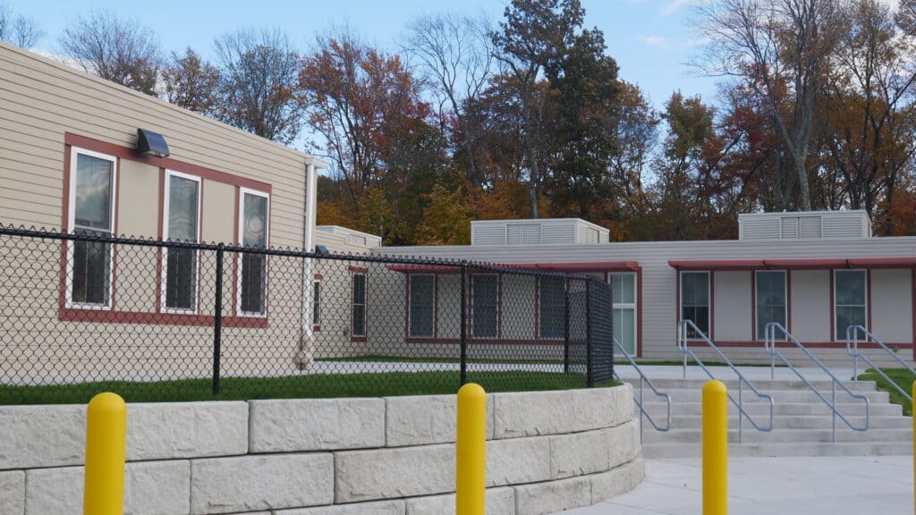 Modern educational facility with a fenced perimeter, featuring a landscaped area and multiple building sections, surrounded by trees in autumn foliage.